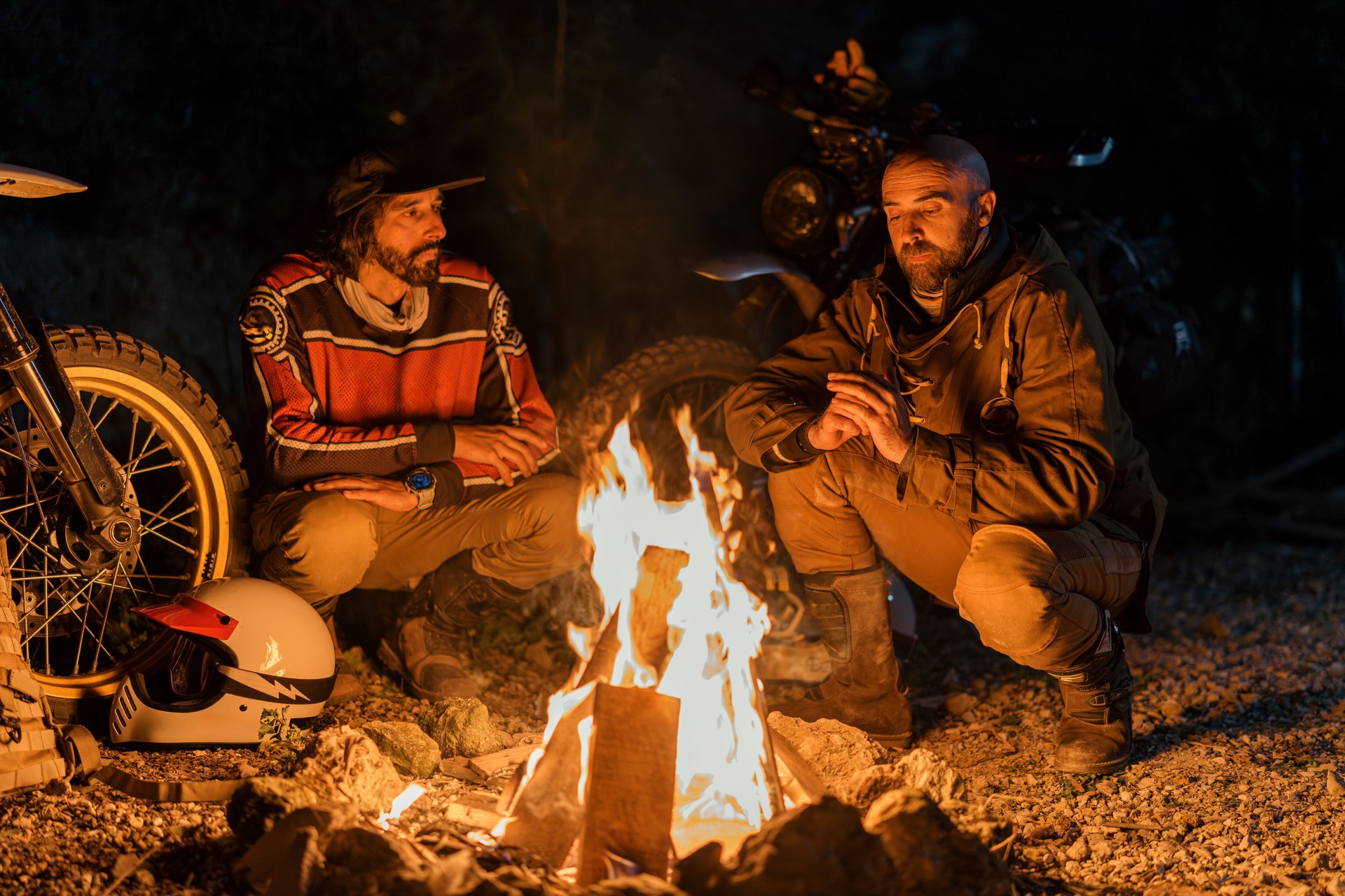 two men kneeling around a campfire at night by their bikes